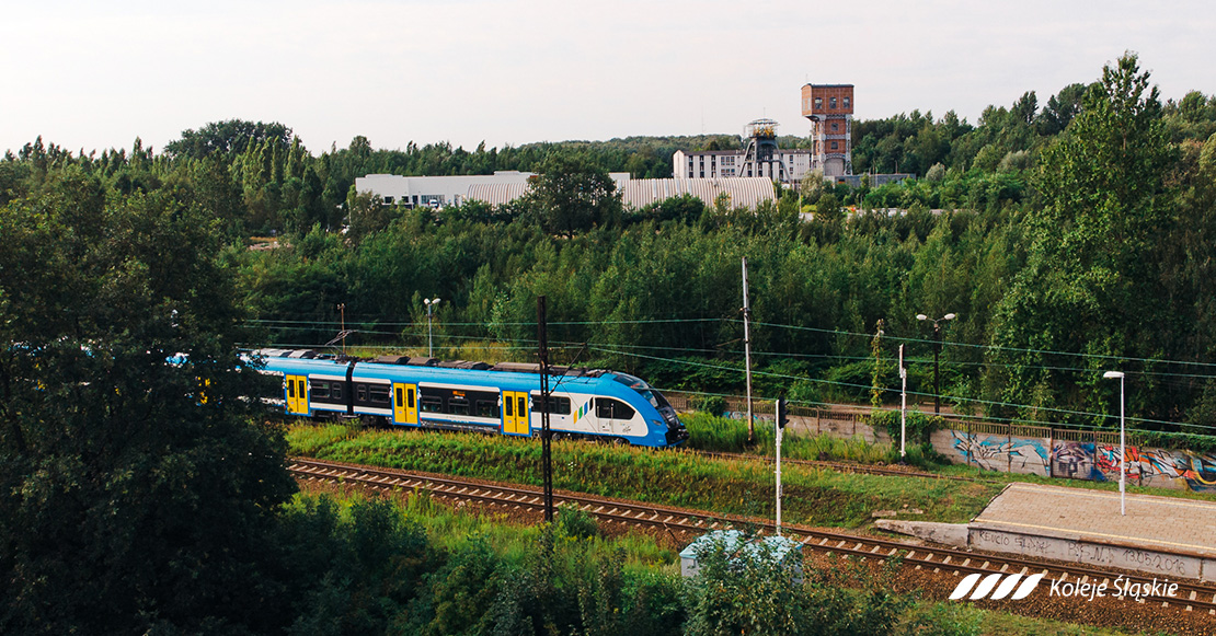 koleje śląskie, pociąg niebiesko-biało-żółty, peron, szyb kopalni na horyzoncie, wśród drzew,